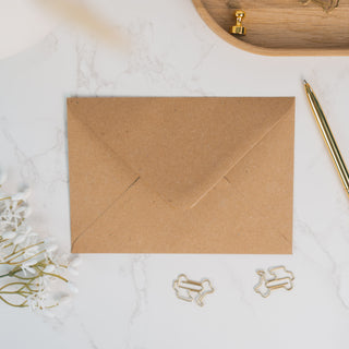 Naturbrauner Briefumschlag im A6-Format, aus recyceltem Kraftpapier, stilvoll arrangiert mit goldenen Büroklammern und einem Kugelschreiber – perfekt für handgeschriebene Botschaften mit einem Hauch von Eleganz.