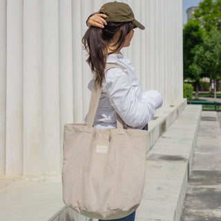 Cordstoff-Tasche Shopper L in der Farbe Beige Delight von De La Mur, getragen von einer Frau mit grüner Kappe in weißer Bluse, vor einer hellen Säulenstruktur.