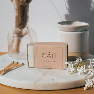 Duftwachswürfel der Sorte Lotusflower & Sandalwood von CAIT Salzburg, präsentiert auf einer Marmorplatte mit dekorativen Elementen und einer Duftwachslampe im Hintergrund.