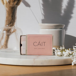 Duftwachswürfel der Sorte Rose Velvet & Precious Oud von CAIT Salzburg, präsentiert auf einer Marmorplatte mit dekorativen Elementen und einer Duftwachslampe im Hintergrund.