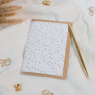 Gratulationskarte mit Konfetti und braunem Umschlag der Marke Typealive liegend auf einem beigen Stoff, dekoriert mit einem goldenen Stift und anderen Accessoires.