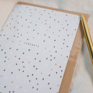 Gratulationskarte mit Konfetti und braunem Umschlag der Marke Typealive liegend auf einem beigen Stoff, dekoriert mit einem goldenen Stift.