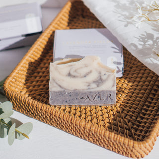 Handgemachte vegane Seife der Sorte Lavender Fields von Azur Natural Bodycare, präsentiert in einem geflochtenen Korb mit ihrer Verpackung im Hintergrund und dekorativen Pflanzen auf einem weißen Untergrund.