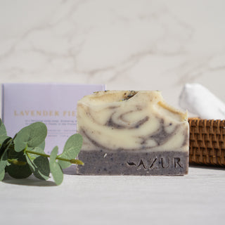 Handgemachte vegane Seife der Sorte Lavender Fields von Azur Natural Bodycare, präsentiert auf einem weißen Untergrund, im Hintergrund ist ein geflochtener Korb mit der Seifenverpackung und dekorativen Pflanzen .