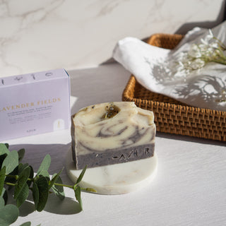 Handgemachte vegane Seife der Sorte Lavender Fields von Azur Natural Bodycare, präsentiert auf einem runde weißen Marmoruntersetzer, im Hintergrund ist ein geflochtener Korb mit der Seifenverpackung und dekorativen Pflanzen sowie ein weißes Tuch.