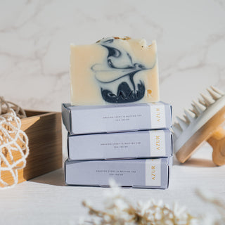 Handgemachte vegane Seife der Sorte Mighty Mint von Azur Natural Bodycare, gestapelt auf ihren Verpackungen mit einer Massagebürste und dekorativen Elementen im Hintergrund auf einem weißen Untergrund.