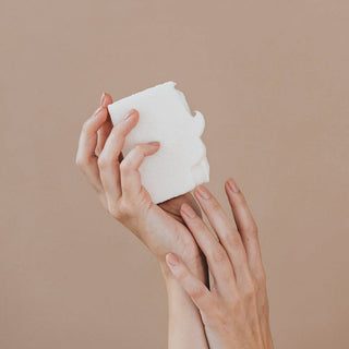 Beiger Hintergrund mit zwei Händen die eine weiße Salzseife der Marke Azur Natural Bodycare hält.