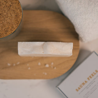 Nahaufnahme einer weißen, duftenden Salzseife aus unserem Sauna Feels Sortiment der Marke Azur Natural Bodycare auf einem Holztablett neben einem weißen Handtuch.