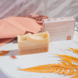 Handgemachte vegane Seife der Sorte Sunset Dreams von Azur Natural Bodycare, präsentiert auf einer Marmorplatte mit ihrer Verpackung und dekorativen Elementen im Hintergrund.