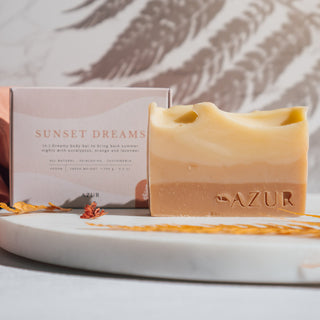 Handgemachte vegane Seife der Sorte Sunset Dreams von Azur Natural Bodycare, präsentiert auf einer Marmorplatte mit ihrer Verpackung und dekorativen Elementen im Hintergrund und einem Schatten.