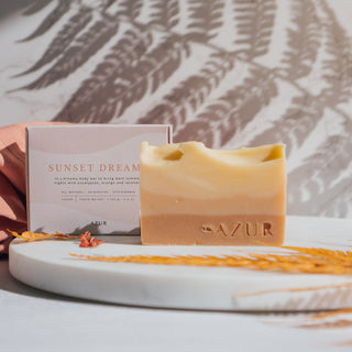 Handgemachte vegane Seife der Sorte Sunset Dreams von Azur Natural Bodycare, präsentiert mit ihrer Verpackung im Hintergrund und dekorativen Elementen auf einer Marmorplatte.
