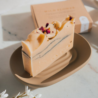 Handgemachte vegane Seife von Azur Natural Bodycare mit dem Namen Wildflower und den Duftnoten von Geranie und Grapefruit, präsentiert auf beiger Seifenablage. Ansicht von oben auf Seife mit getrockneten Blütenblätter.