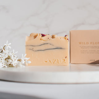 Handgemachte vegane Seife von Azur Natural Bodycare mit dem Namen Wildflower und den Duftnoten von Geranie und Grapefruit, präsentiert mit eleganter Verpackung auf einer Marmorfläche und weißen Blumen.