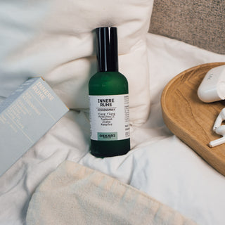Innere Ruhe Kissenspray der Marke Oskari Berlin, präsentiert auf einem Bett mit weißem Kissen, eine hellbaue Verpackung aus Karton des Produktes, sowie eine Schlafmaske und Holztablett.