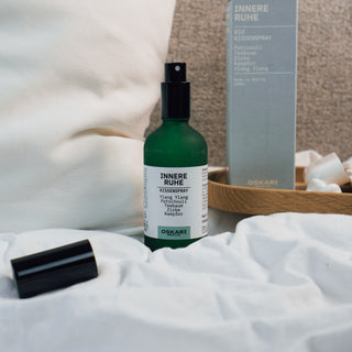Innere Ruhe Kissenspray der Marke Oskari Berlin, präsentiert auf einem Bett mit weißem Kissen, im Hintergrund eine hellbaue Verpackung aus Karton des Produktes.
