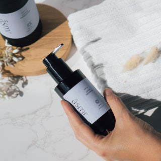 Körper- und Massageöl der Sorte Lotus und Geranie von Asmi Ayurveda, die Flasche wird von einer Hand gehalten und im Hintergrund ist ein weißes Handtuch zu sehen.
