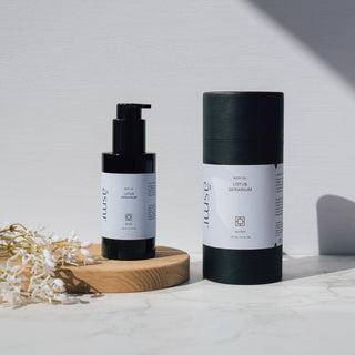 Körper- und Massageöl der Sorte Lotus und Geranie von Asmi Ayurveda, präsentiert in einer schwarzen Flasche und Verpackung auf einem Holztablett und Marmortisch mit dekorativen Blumen.