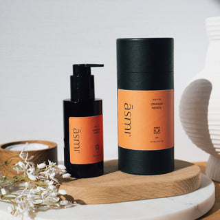 Körperöl der Sorte Orange und Neroli von Asmi Ayurveda, präsentiert in einer schwarzen Flasche und Verpackung auf einem Holztablett und Marmortisch mit dekorativen Blumen und einer Kerze.