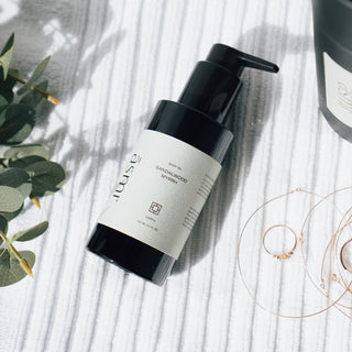 Körperöl der Sorte Sandelholz und Myrrhe von Asmi Ayurveda, liegend auf einem weißen Handtuch mit dekorativen grünen Blättern im Hintergrund und Schmuck.