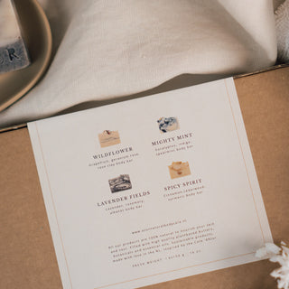 Die 'Le Quartet' Geschenkbox von Azur Natural Bodycare, eine umweltfreundliche Verpackung aus braunem Kraftpapier mit einem eleganten weißen Label und minimalistischer Schrift. Die Szene ist auf auf beigem Stoff, einer Seife und mit Blumen gestaltet, was eine schlichte, aber stilvolle Ästhetik vermittelt.
