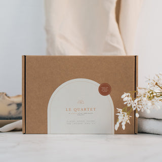 Die 'Le Quartet' Geschenkbox von Azur Natural Bodycare, eine umweltfreundliche Verpackung aus braunem Kraftpapier mit einem eleganten weißen Label und minimalistischer Schrift. Die Szene ist auf einer weißen Marmoroberfläche mit beigem Stoff im HIntergrund und Blumen gestaltet, was eine schlichte, aber stilvolle Ästhetik vermittelt.