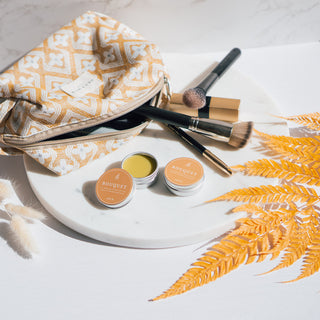 Handgemachter veganer Lippenbalsam der Sorte Bouquet von Azur Natural Bodycare, arrangiert mit einer gelben Kosmetiktasche, Make-up-Pinseln und orangene Blättern auf einer dekorativen Marmorplatte.