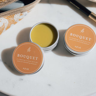 Handgemachter veganer Lippenbalsam der Sorte Bouquet von Azur Natural Bodycare, arrangiert auf einer Marmorplatte mit einer gelben Kosmetiktasche und schwarzem Eyelinerstift.