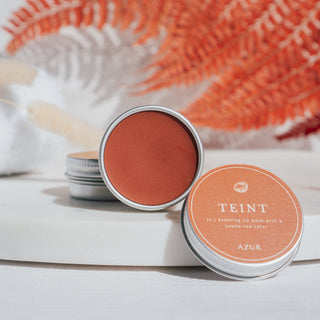 Handgemachter veganer Lippenbalsam der Sorte Teint von Azur Natural Bodycare, präsentiert auf einer Marmorplatte mit dekorativen roten Blättern und einem weißen Handtuch im Hintergrund.