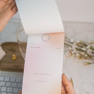 Eine Hand blättert ein Blatt des Notizblocks „Daily Tasks“ von Typealive um. Der elegante Planer mit sanftem Farbverlauf in Rosa und Beige bietet eine strukturierte Übersicht für tägliche Aufgaben, Affirmationen und Dankbarkeitsmomente. Im Hintergrund sorgen weiße Blüten und ein minimalistisches Schreibtisch-Setting für eine inspirierende Atmosphäre. Perfekt für produktive Tage mit Stil.