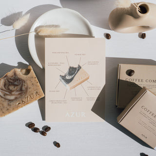 Handgemachte vegane Peelingseife der Sorte Coffee Comfort von Azur Natural Bodycare, präsentiert mit einem Infoflyer über die Seife, Kaffeebohnen, einem Handtuch und einem Keramikgefäß auf einem weißen Untergrund.