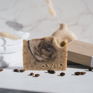 Handgemachte vegane Peelingseife der Sorte Coffee Comfort von Azur Natural Bodycare, präsentiert mit Kaffeebohnen, einem Handtuch und einem Keramikgefäß auf einem weißen Hintergrund.