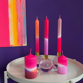Tisch mit bunten pinken Stabkerzen und Stumpenkerzen der Marke Pink Stories, im Hintergrund ist eine lila Wand und ein buntes Bild zu sehen.