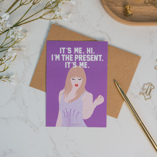 Lila Postkarte mit dem Spruch “It’s Me. Hi. I’m the Present. It’s Me.”, illustriert mit einer stilisierten Frau in glitzerndem Kleid, inklusive braunem Kraftpapier-Umschlag – eine originelle Geburtstagskarte für Musikfans.