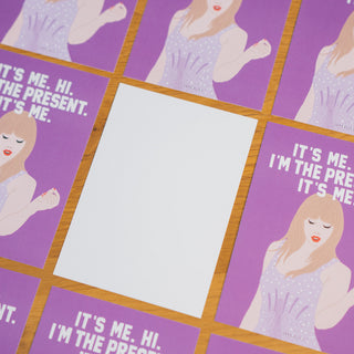 Die Postkarte “It’s Me. Hi. I’m the Present. It’s Me.” von Typealive liegt auf einem Holztisch, umgeben von mehreren identischen Karten. Eine Postkarte ist umgedreht, sodass die weiße Rückseite sichtbar ist – perfekt zum individuellen Beschriften für persönliche Nachrichten.