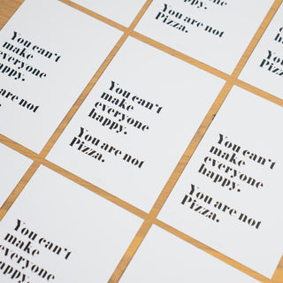 Mehrere Postkarten mit dem humorvollen Spruch ‘You can’t make everyone happy. You are not Pizza.’ in schwarzer Schrift auf weißem Hintergrund, stilvoll auf einem Holztisch angeordnet von Typealive.