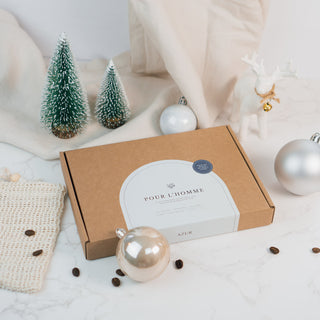 Die 'Pour L’Homme' Geschenkbox von Azur Natural Bodycare, eine umweltfreundliche Verpackung aus braunem Kraftpapier mit einem eleganten weißen Label und minimalistischer Schrift. Im Vordergrund liegt ein kunstvoll gemustertes Seifenstück auf einem natürlichen Netz-Seifenbeutel, umgeben von verstreuten Kaffeebohnen, die das Pflegeprodukt thematisch ergänzen. Die Szene ist auf einer weißen Marmoroberfläche mit beigem Stoff und dezenten Weihnachtsdekoration gestaltet, was eine schlichte, aber stilvolle Ästheti