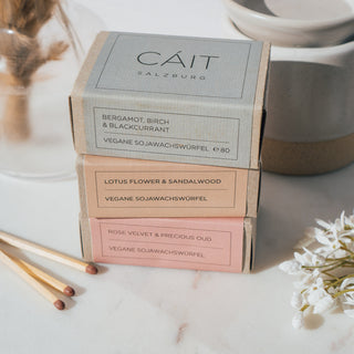 Produktübersicht der Duftwachswürfel von CAIT Salzburg in den Sorten Bergamot, Birch & Blackcurrant, Lotus Flower & Sandalwood, und Rose Velvet & Precious Oud, präsentiert auf einer Marmorplatte mit dekorativen Elementen und Streichhölzern.