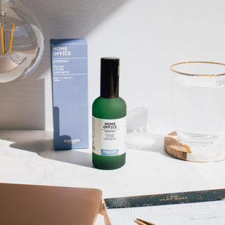 Raumspray Home Office in der Duftrichtung Orange Litsea Lemongrass von Oskari Berlin, abgebildet neben der Verpackung, einer Glühbirne und einem Glas im Hintergrund.