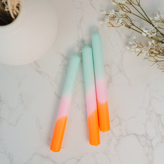 Drei handgefärbte Stabkerzen aus dem Set „Dip Dye Neon – Frühlingssorbet“ von Pink Stories liegen auf einer Marmorfläche. Die Kerzen haben einen sanften Farbverlauf von Mintgrün über Rosé bis hin zu leuchtendem Neonorange. Im Hintergrund sind eine schlichte Vase und weiße Blütenzweige zu sehen.