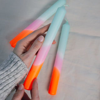 Eine Hand hält eine der handgefärbten Stabkerzen aus dem Set „Dip Dye Neon – Frühlingssorbet“ von Pink Stories. Die Kerze zeigt einen sanften Farbverlauf von Mintgrün über Rosé bis zu intensivem Neonorange. Im Hintergrund liegen weitere Kerzen auf einem beigen Stoff, was die weiche und moderne Ästhetik des Produkts betont.