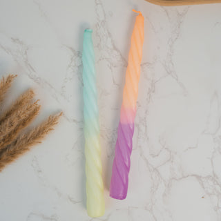 Zwei gedrehte Stabkerzen aus dem Set „Rainbow Studio – Fancy“ von Pink Stories liegen auf einer Marmorfläche. Die eine Kerze zeigt einen sanften Farbverlauf von Mintgrün zu Pastellgelb, während die andere von leuchtendem Orange in ein kräftiges Violett übergeht. Links im Bild liegt dekoratives Pampasgras.