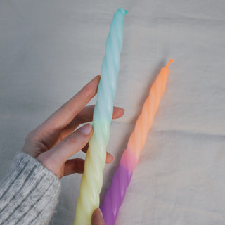 Eine Hand hält eine der zwei gedrehten Stabkerzen aus dem Set „Rainbow Studio – Fancy“ von Pink Stories. Die Kerze hat einen Farbverlauf von Mintgrün zu zartem Gelb. Im Hintergrund liegt die zweite Kerze mit einem sanften Übergang von leuchtendem Orange zu kräftigem Violett auf beigem Stoff.