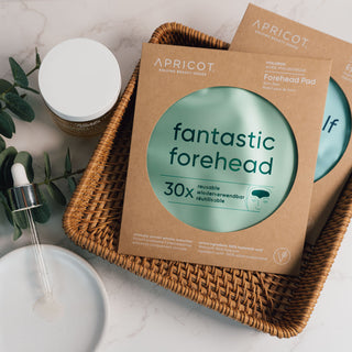 Wieverwendbare Stirnpads mit Hyaluron der Marke Apricot Beauty & Healthcare in umweltfreundlicher Kartonverpackung, liegend präsentiert auf einer marmorierten Oberfläche und dekoriert mit Eukalyptus und geflochtenem Korb.