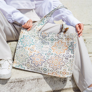 Stofftasche Everyday Bag M im Muster Floral Harmony von De La Mur, getragen von einer sitzenden Person in heller Kleidung und weißen Sneakern, vor einer hellen Struktur.