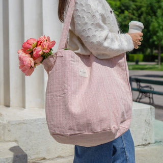 Frau trägt eine Stofftasche Shopper L in der Farbe Blushing Pink von De La Mur, mit Blumen gefüllt, im Freien vor einer Säule und hält eine Tasse Kaffee.