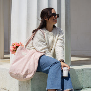 Stofftasche Shopper L in der Farbe Blushing Pink von De La Mur, getragen von einer Frau in cremefarbenem Pullover und blauen Jeans, die einen Kaffee hält und auf einer hellen Stufe sitzt, vor einer Säulenstruktur.