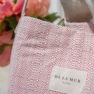 Nahaufnahme der Stofftasche Shopper L in der Farbe Blushing Pink von De La Mur, zeigt das gewebte Muster und das Markenlabel vor einem Hintergrund aus rosa Blumen.