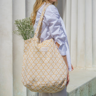 Frau trägt eine Stofftasche Shopper L in der Farbe Sunny Geometric von De La Mur, gefüllt mit weißen Blumen, im Freien vor einer Säule.