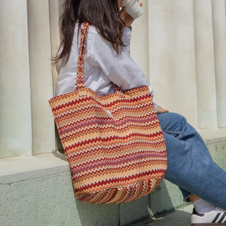 Frau sitzt im Freien vor einer Säule und trägt eine Stofftasche Shopper L in der Farbe Zigzag Sunset von De La Mur, mit einer Tasse Kaffee in der Hand.