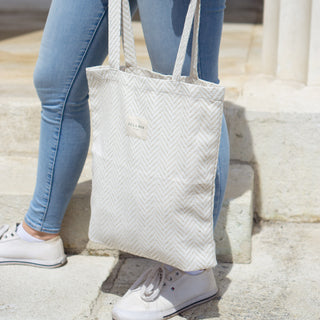 Frau hält eine Stofftasche Shopper M in der Farbe Chevron Cream von De La Mur, während sie in Jeans und weißen Sneakern vor einer Säule steht.